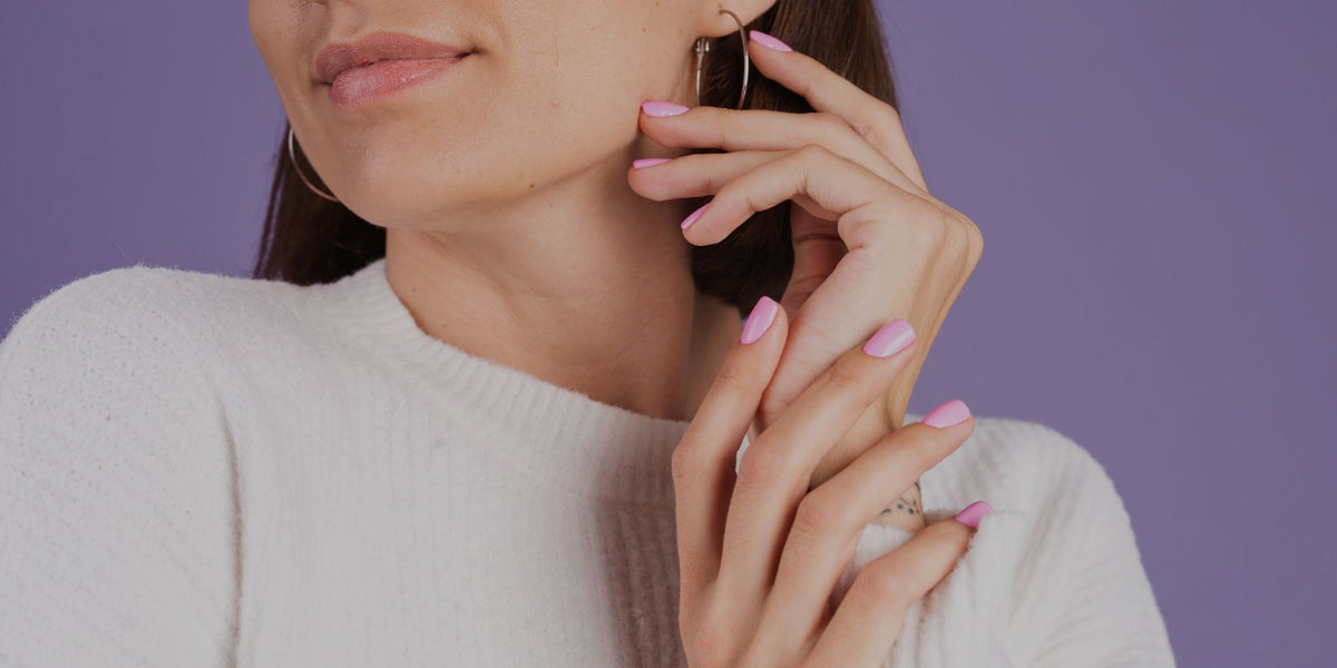 A close-up lifestyle shot of a woman wearing gold hoop earrings, a ring, and pink manicured nails, wearing a white knit sweater against a solid purple background.
