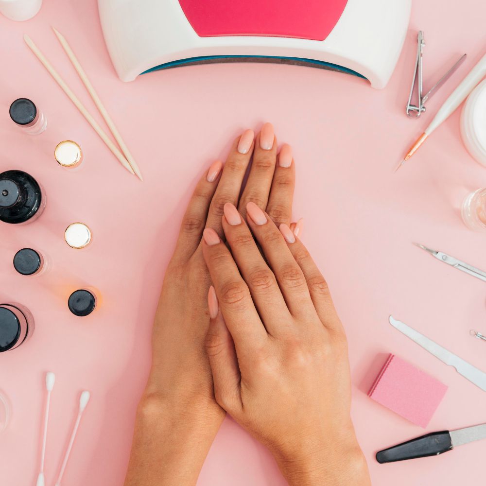 A top-down view of two hands with neatly manicured pink nails resting on a pink spa table, surrounded by nail care tools and bottles including a UV lamp, cuticle oil, cotton swabs, and nail files.
