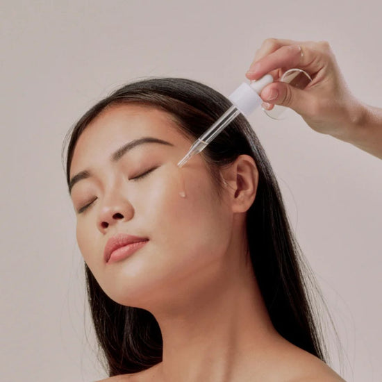 Person applying a dropper of skincare product to their face against a neutral background
