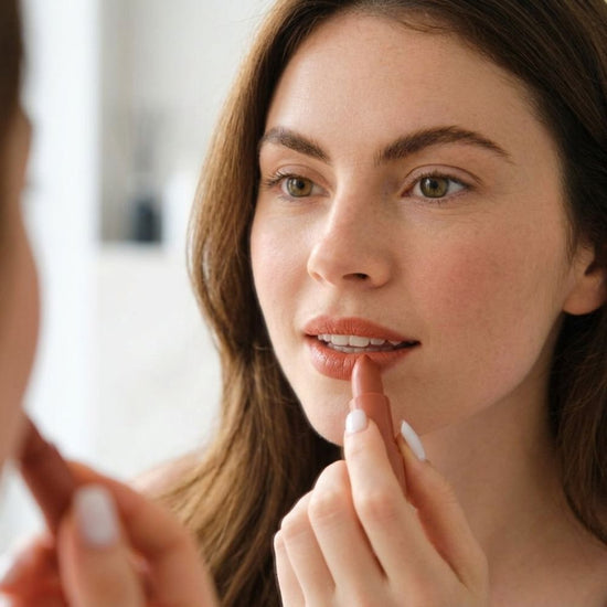 A close-up lifestyle shot of a woman applying a nude lip color from a lip gloss tube while looking into a mirror.