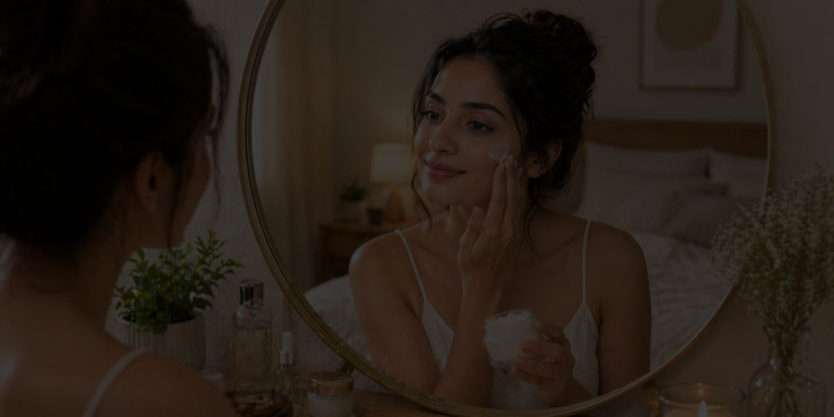 A woman applies skincare cream to her face while looking at her reflection in a round mirror in a warmly lit bedroom.