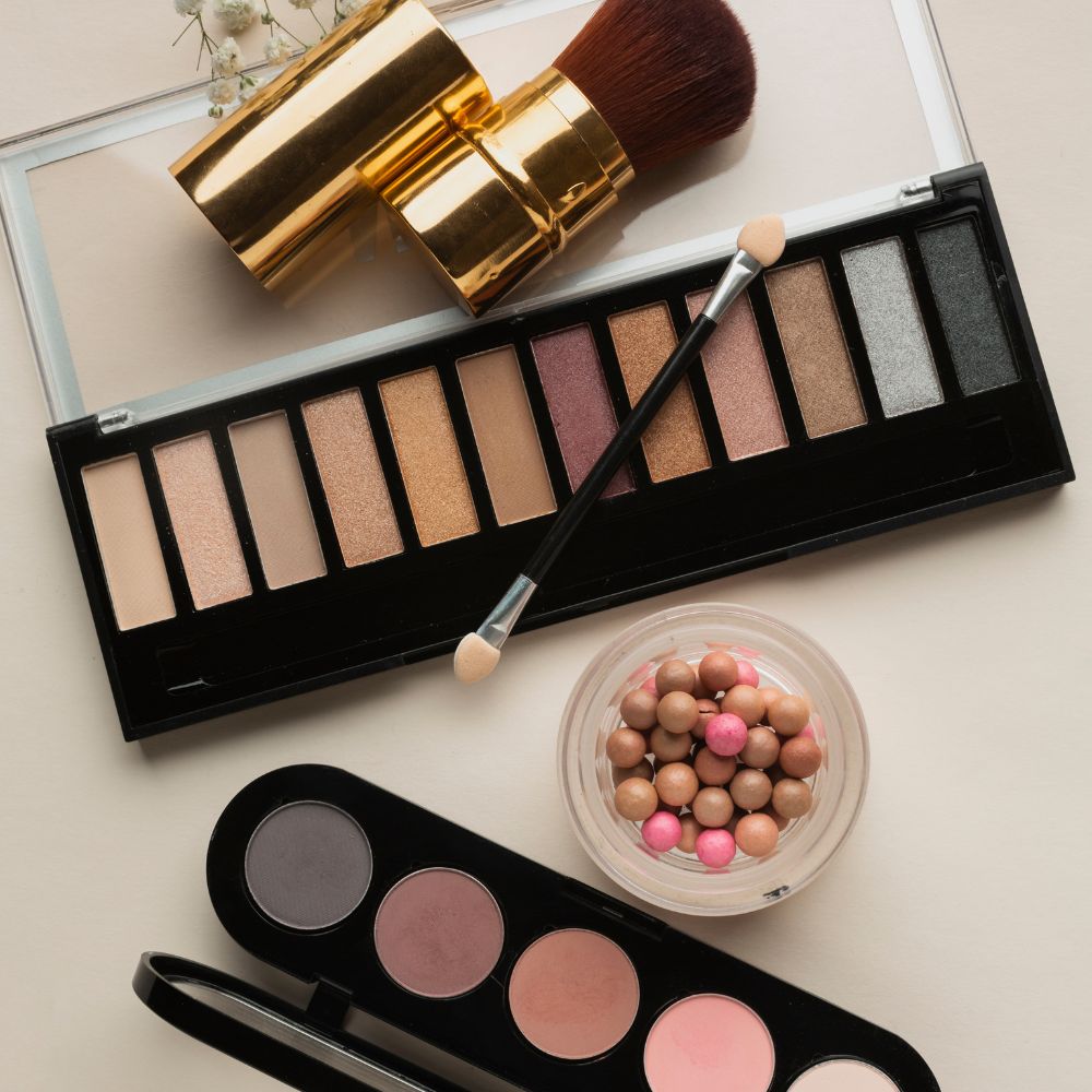 An open makeup palette with multiple eyeshadow pans, a small jar of round makeup beads, and a makeup brush arranged on a light beige background.