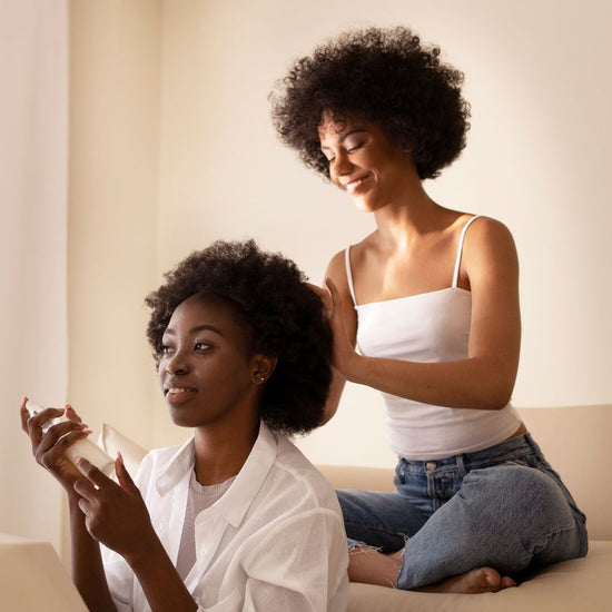 Two women in a warm, softly lit indoor setting. One woman with dark curly hair sits holding a small clear bottle, while another woman with dark curly hair sits behind her, placing her hand on her head.