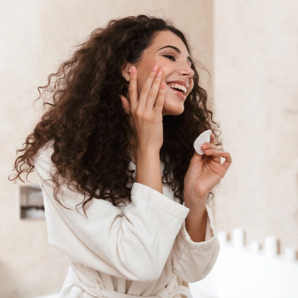 A woman with long, curly brown hair wearing a white bathrobe smiles while applying or spreading a white cosmetic cream with a small white spatula in a bright bathroom setting.