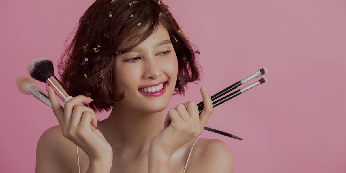 A smiling woman with short brown hair holds makeup brushes against a solid pink background.
